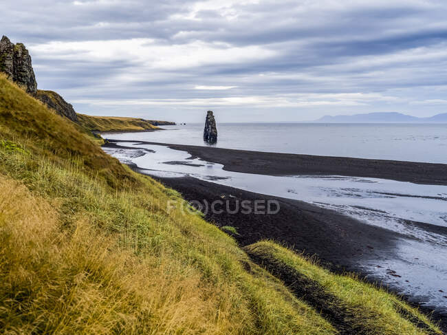 Tall rock formation and grassy slopes along the shoreline of a fjord; Iceland — Stock Photo