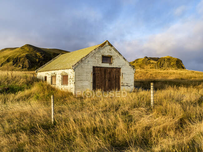 Выветренный сарай в травянистой зоне с проволочным забором и скалистыми обнажениями на заднем плане; Исландия — стоковое фото