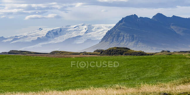 Paysage dans le sud-est de l'Islande avec des montagnes accidentées et un champ d'herbe, et des montagnes enneigées au loin ; Hornafjorour, région orientale, Islande — Photo de stock