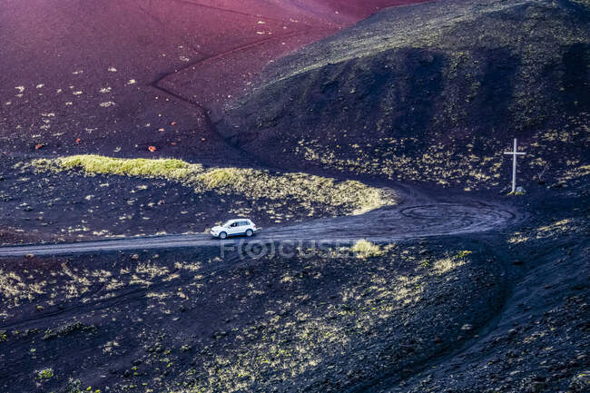 Coche que viaja en una carretera en el paisaje volcánico negro con una cruz en el borde de la carretera en una de las islas Westman; Vestmannaeyjar, Región del Sur, Islandia - foto de stock