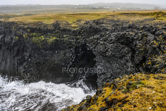 View point from a cliff to the waves rolling into a volcanic cave along the coastline in Arnarstapi; Snaefellsbaer, Western Region, Iceland — стоковое фото