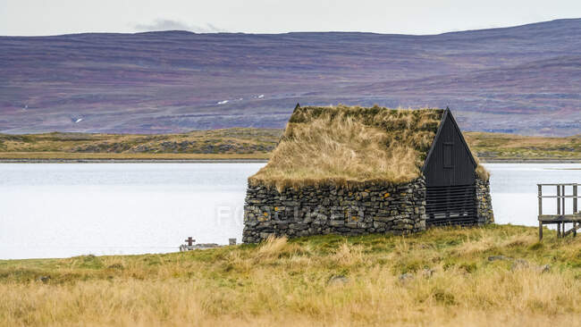 Steingebäude mit Grasdach zum Fjord und einem Kreuz im Gras davor; Sudavik, Westfjord, Island — Stockfoto
