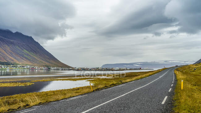 Дорога вдоль спокойной воды фьорда со зданиями поселка Isafjorour на другой стороне; Isafjardarbaer, Westfjords, Исландия — стоковое фото