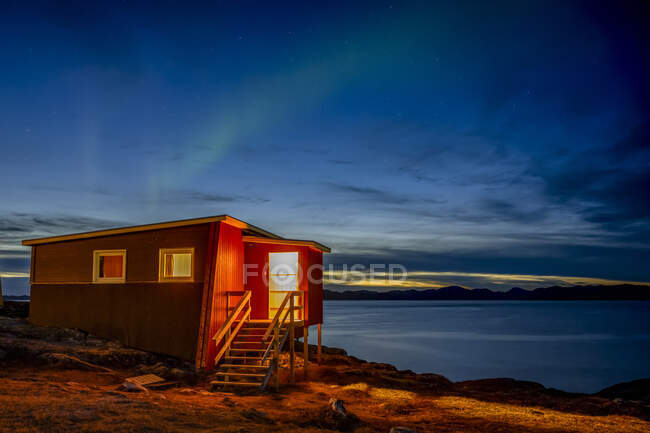 Небольшой дом на краю воды с видом на спокойную береговую линию в ночное время и мерцающее северное сияние сверху; Нуук, Сермерсук, Гренландия — стоковое фото