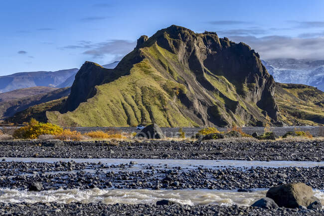 Formación de tierras robustas cubiertas de follaje verde con agua que fluye en primer plano; Rangarthing eystra, Región Sur, Islandia - foto de stock