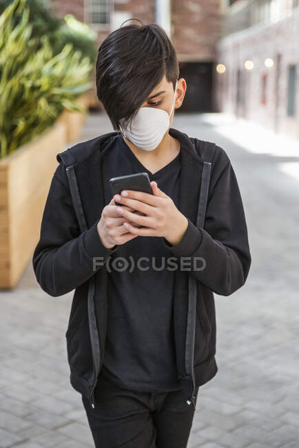 Niño usando su teléfono inteligente y usando una máscara protectora para proteger contra COVID-19 durante la Pandemia Mundial del Coronavirus; Toronto, Ontario, Canadá - foto de stock