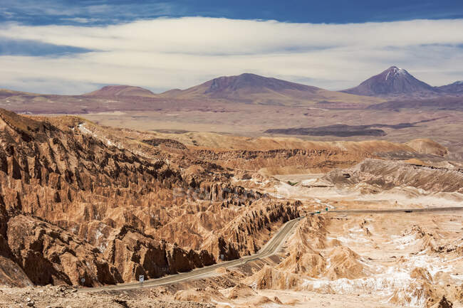 Route descend dans une vallée désertique de haute altitude avec des formations rocheuses uniques sur la gauche, et un pic volcanique au loin ; San Pedro de Atacama, Antofagasta, Chili — Photo de stock