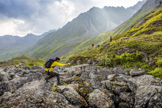 A caucasian male jumps from boulder to boulder across a boulder field, with an asian man and caucasian woman and their dog hike in the background on the very rocky/bouldery Reed Lakes trail through the Talkeetna Mountains in Hatcher's Pass, Palmer, — Stock Photo