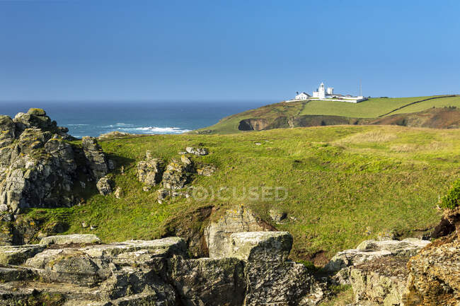 Farol branco no topo de campos verdes montanhosos emoldurados por afloramentos rochosos e céu azul; Condado de Cornwall, Inglaterra — Fotografia de Stock