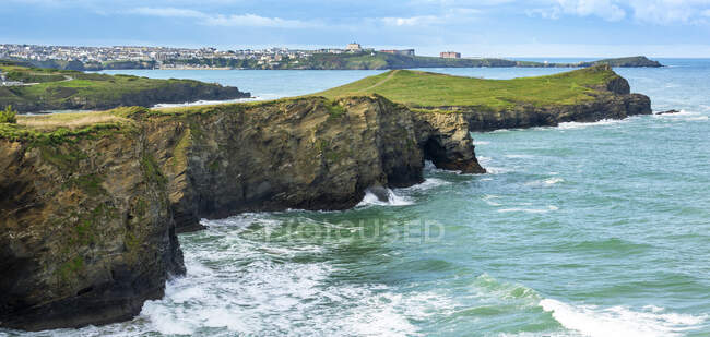 Rocky cliff shoreline peninsulas with green fields, blue sky and town in the far background; Cornwall County, England — Stock Photo