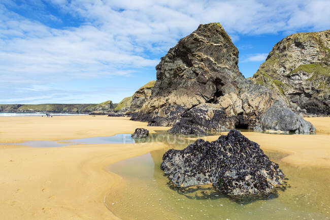 Plage de sable avec des formations rocheuses le long d'une falaise rocheuse rivage avec surf et ciel bleu et nuages ; comté de Cornwall, Angleterre — Photo de stock