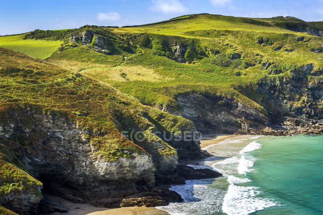 Acantilados rocosos a lo largo de la costa con colinas cubiertas de hierba, cielo azul y nubes; Condado de Cornwall, Inglaterra - foto de stock