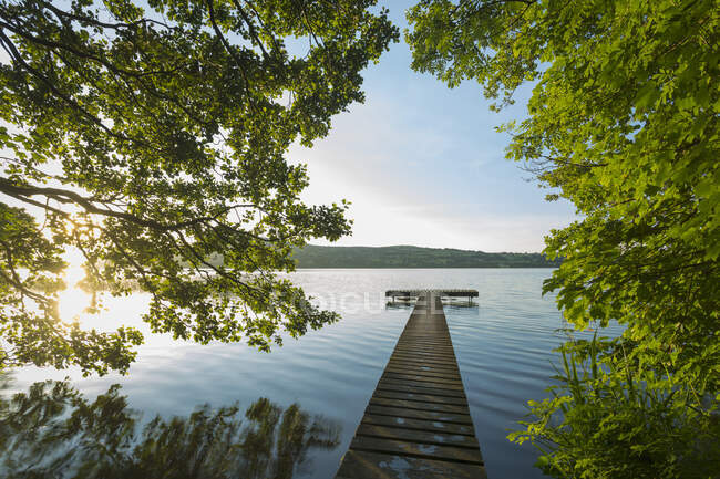 Wooden pier of the banks of Lough Derg lake framed by the surrounding trees, with the rising sun glistening through the leaves; Killaloe, County Clare, Ireland — Stock Photo