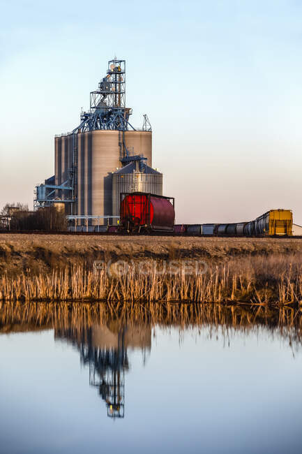 Instalação de armazenamento de grãos e trem nas pradarias de Alberta refletidas na água; Alberta, Canadá — Fotografia de Stock