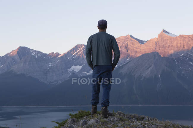 Hombre de pie en una cresta con vistas a un lago hacia los picos rocosos de la montaña; Kananaskis, Alberta, Canadá - foto de stock