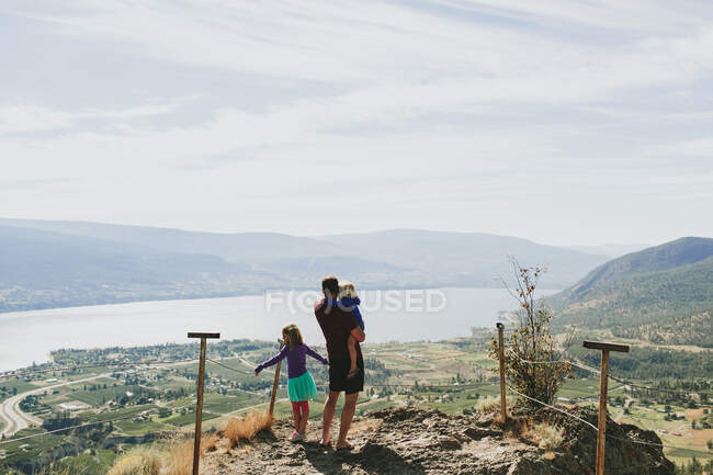 Un père avec ses filles debout sur un bord de rocher surplombant le lac Okanagan ; Peachland, Colombie-Britannique, Canada — Photo de stock