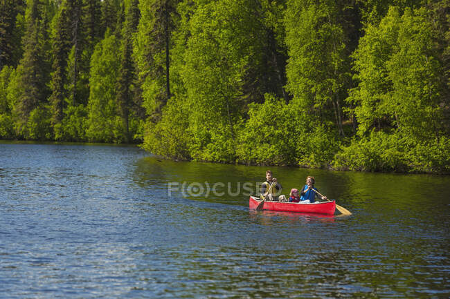 Una pareja y una joven en una canoa roja en el lago Byers con costa verde boscosa en el campamento del lago Byers, Denali State Park; Alaska, Estados Unidos de América - foto de stock
