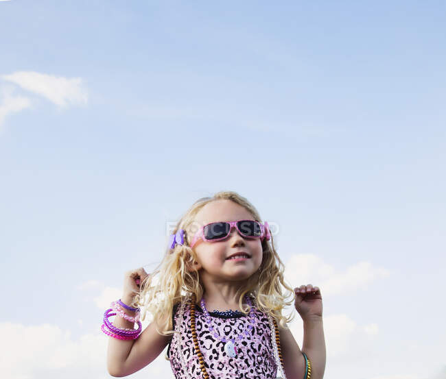 Chica con el pelo rubio rizado con gafas de sol y joyas de pie haciendo caras lindas contra un cielo azul con nubes; Spruce Grove, Alberta, Canadá - foto de stock