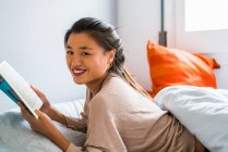 Joven atractivo asiático mujer leyendo libro en cama - foto de stock