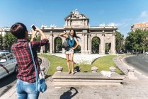 Chinese female tourist taking shot of Japanese friend on background of Puerta de Alcala (Alcala Door) in Madrid, Spain. — Stock Photo