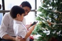 Азіатська сім'я святкує Різдво, хлопчик використовує смартфон біля ялинки — стокове фото