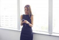 Молодая азиатская деловая женщина, использующая смартфон за окном в современном офисе — стоковое фото