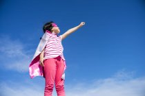Cute little superhero girl in mask and coat posing against sky — Stock Photo