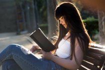 Junge eurasische Frau liest ein Buch auf Bank im Park — Stockfoto