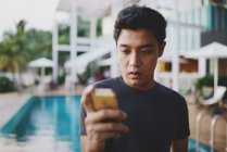 Giovane attraente asiatico utilizzando smartphone contro piscina — Foto stock