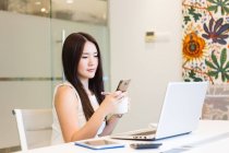 Jeune femme regardant son téléphone dans une salle de réunion, assise devant son ordinateur portable . — Photo de stock