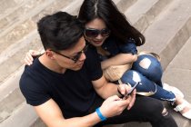 Beautiful young asian couple sitting on steps with smartphone — Stock Photo