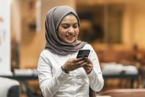 Молода азіатська бізнес-леді в хіджабі, використовуючи смартфон в сучасному офісі — стокове фото
