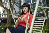 Young lady using cellphone on swig in the park, Singapore — Stock Photo