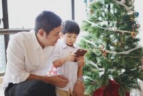 Asian family celebrating Christmas holiday, father and son with smartphone near fir tree — Stock Photo