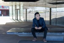 Молодий азіатський чоловік сидить у навушниках на вулиці — стокове фото