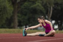 A young asian female is stretching herself on a race track in bad weather. She is preparing herself for the daily exercise, despite the weather. — Stock Photo