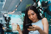 Молода азіатська жінка використовує смартфон і має напій — стокове фото