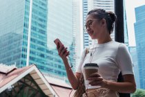 Молода приваблива азіатська жінка використовує смартфон і тримає каву — стокове фото