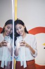 Junge Asiatin steht vor dem Spiegel und benutzt Smartphone — Stockfoto