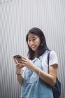 Молодой студент азиатского колледжа, использующий смартфон против серой стены — стоковое фото