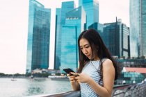Jovem asiático mulher usando smartphone contra arranha-céus — Fotografia de Stock