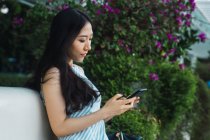 Вид збоку молодої азіатки, що використовує смартфон проти квітів — стокове фото