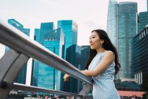 Junge Asiatin steht auf Geländer mit Wolkenkratzern im Hintergrund — Stockfoto