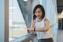Молода азіатська бізнес-леді розмовляє по смартфону і перевіряє час в сучасному офісі — стокове фото