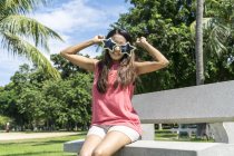 Щаслива азіатська доросла жінка сидить на лавці і позує в смішних сонцезахисних окулярах — стокове фото