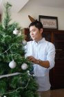 Happy young asian man decorating fir tree — Stock Photo