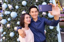 Jeune attrayant asiatique couple ensemble shopping dans le centre commercial à Noël et de prendre selfie contre sapin — Photo de stock