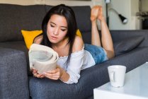 Joven mujer china y bonita leyendo un libro en el sofá - foto de stock