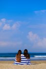 Дві подруги-жінки з довгим волоссям сидять на пляжі у синьому та білому рушнику, насолоджуючись видом на океан . — стокове фото