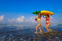 Dos chicas guapas con flotadores camino a las olas del océano . - foto de stock